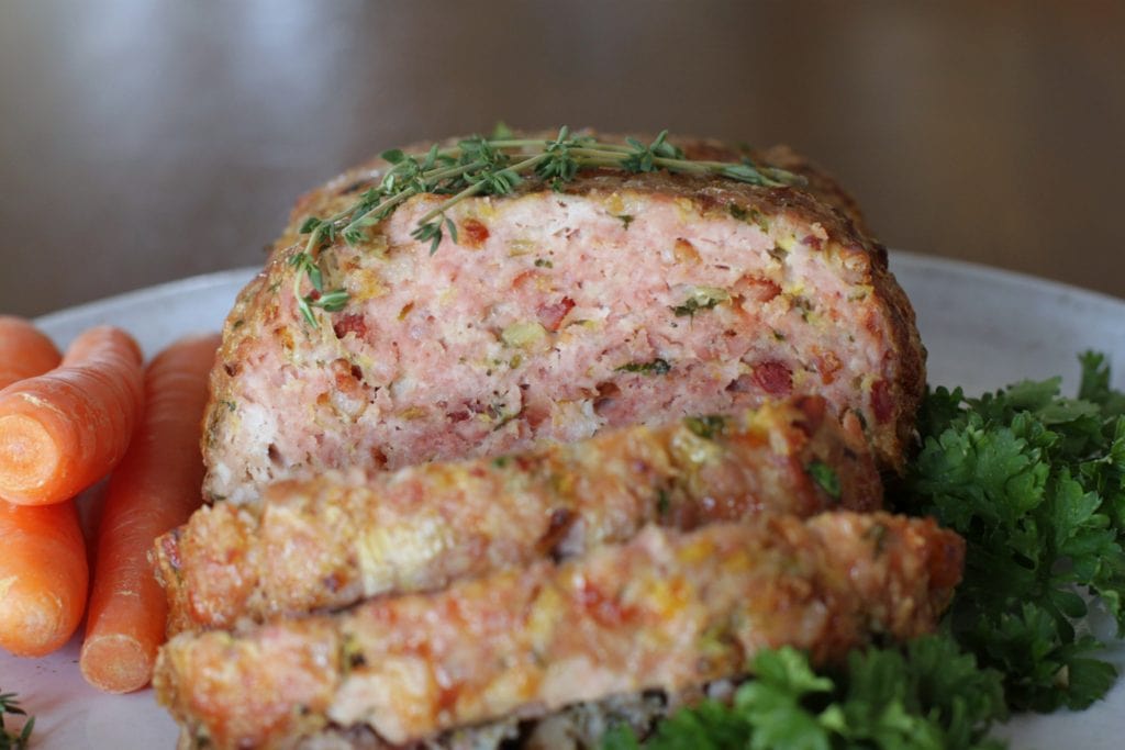 Close up of a sliced pork meatloaf topped with thyme and surrounded by carrots and kale.